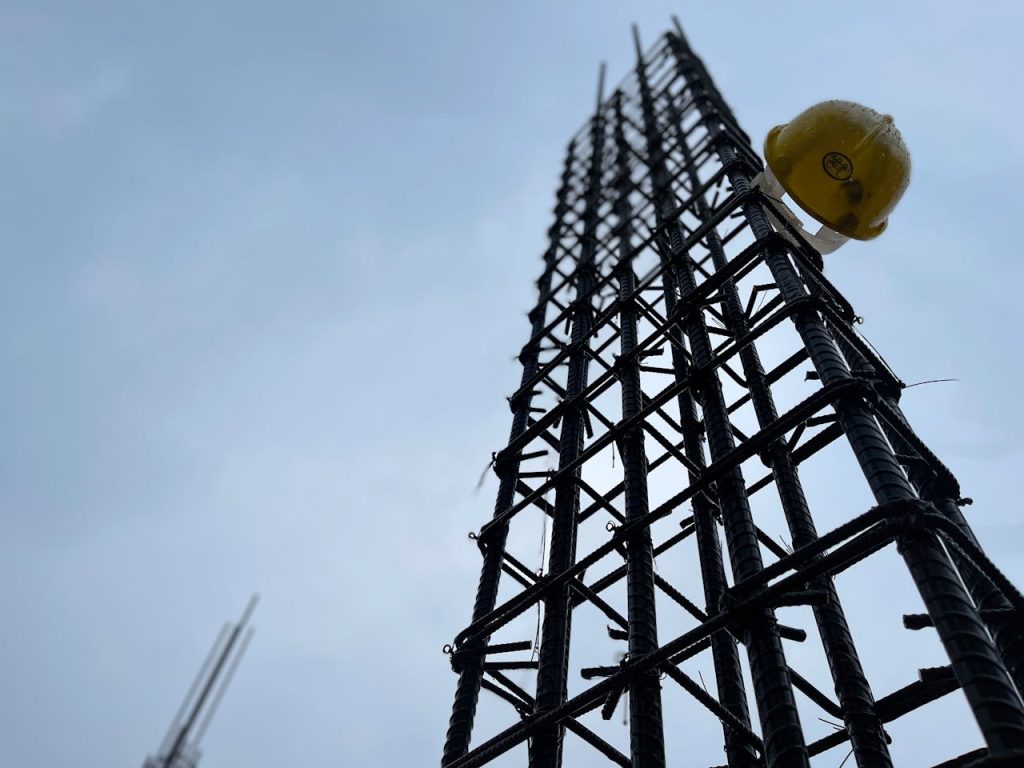 Vertical steel reinforcement bars on site with hanging helmet indicating construction safety focus.